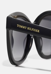 Black oversized sunglasses with gradient lenses and a sleek, glossy frame. The side features gold "TOMMY HILFIGER" branding.