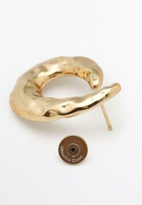 Náušnice z kovu se zlatým tónem s vlnitým, nepravidelným designem, vybavené zapínáním na puzetu a malým kruhovým logem. Hladká, lesklá textura.