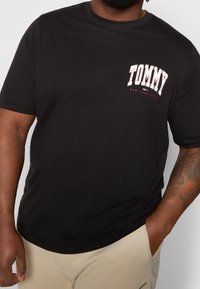Camiseta de algodón negra con la palabra "TOMMY" en blanco y con acentos rojos, de mangas cortas y diseño de cuello redondo. Textura suave.