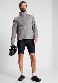 Veste de cyclisme grise légère avec fermeture éclair à l'avant et poches, associée à un short de cyclisme noir rembourré. L'athlète tient un casque noir.