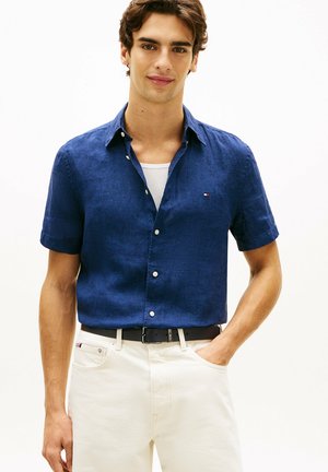 Jonge man die een navyblauw overhemd met korte mouwen draagt over een wit ondershirt en witte broek met een zwarte riem, hand in de zak.