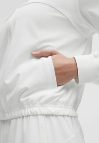 Vit plagg med en mjuk, slät textur; har en sidoficka och en rynkad elastisk midja. Ärmarna har en ren avslutning.