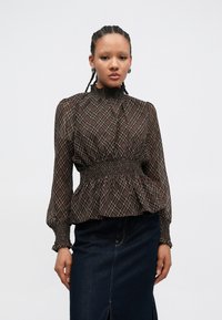 Brun bluse med mønster, høy smokkekrage og elastisk midje, kombinert med en mørk denimskjørt. Lange ermer med smokkede mansjetter.