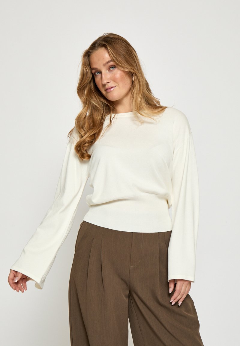 Biele dlhé rukávy top s uvoľneným strihom a okrúhlym výstrihom, skombinovaný s vysokými hnedými skladanými nohavicami. Mäkká, hladká textúra látky.