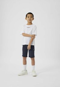 Hvit bomulls t-skjorte med svart tekst, kombinert med marineblå shorts. Hvite joggesko og hvite sokker. Barn står med armene krysset, nøytral bakgrunn.