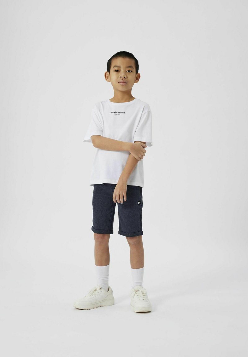 Hvit bomulls t-skjorte med svart tekst, kombinert med marineblå shorts. Hvite joggesko og hvite sokker. Barn står med armene krysset, nøytral bakgrunn.