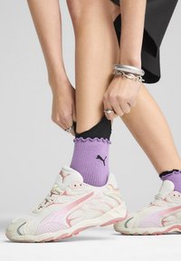 Zapatillas deportivas blancas y rosas con parte superior de malla, diseño curvado, suela de goma, acompañadas de calcetines morados con bordes rizados y acentos negros.
