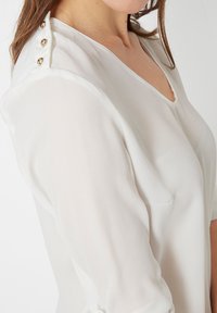 Mujer vestida con una blusa blanca con botones a presión dorados en el hombro, que muestra un escote en V y mangas largas sobre un fondo liso.