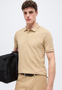 Jeune homme portant un polo beige et un pantalon, tenant un sac noir à bandoulière sur l'épaule, debout devant un fond blanc uni.