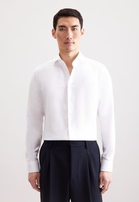 Camicia bianca a maniche lunghe con colletto alla coreana, dotata di orlo dritto e chiusura a bottoni, abbinata a pantaloni scuri aderenti.