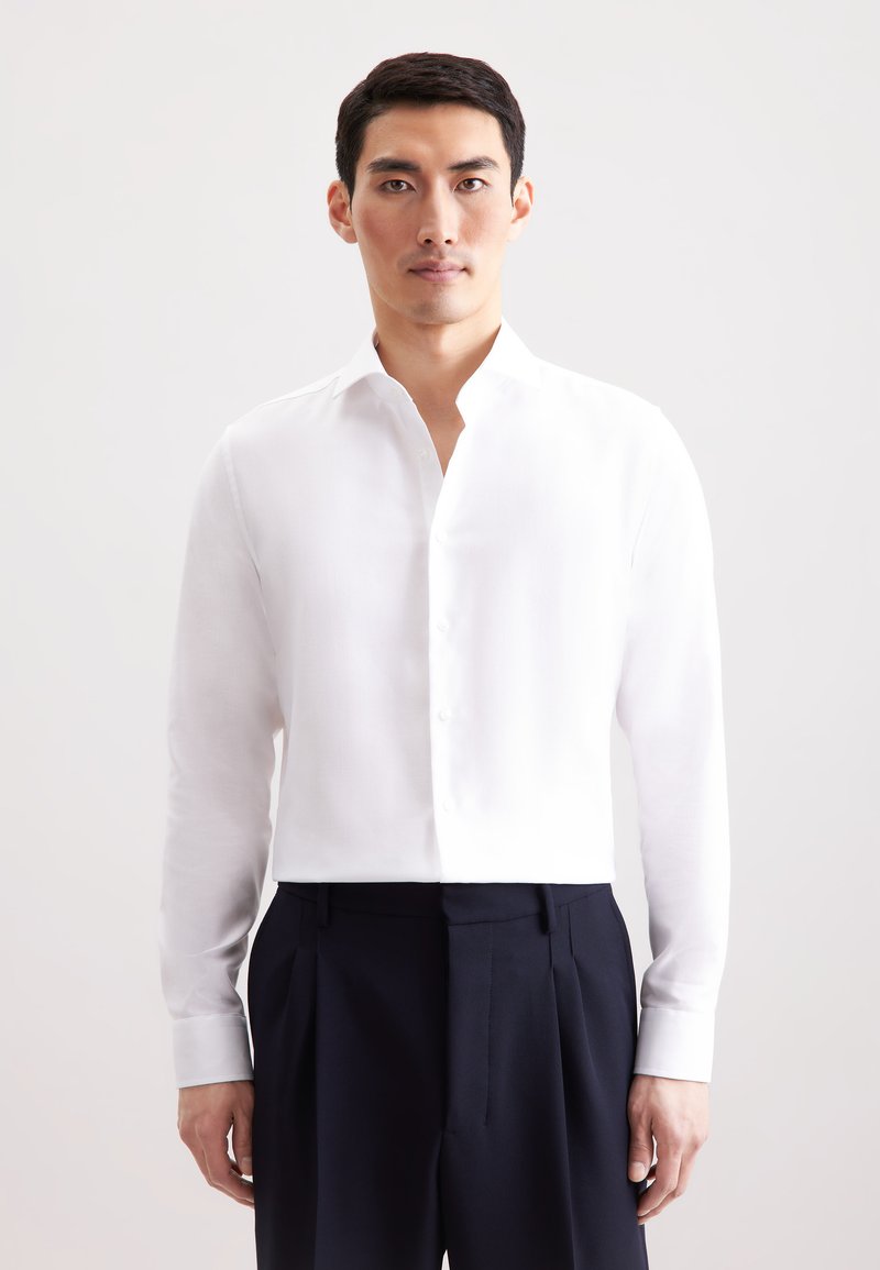 Camicia bianca a maniche lunghe con colletto alla coreana, dotata di orlo dritto e chiusura a bottoni, abbinata a pantaloni scuri aderenti.