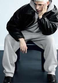 Čierna kožená oversize bunda cez šedú mikinu a tepláky. Subjekt sedí na modrej stoličke, s hladkým, minimalistickým pozadím.