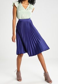 Gonna plissè fino al ginocchio di un profondo viola, realizzata in tessuto leggero. Abbinata a una blusa verde chiaro con maniche a volant.