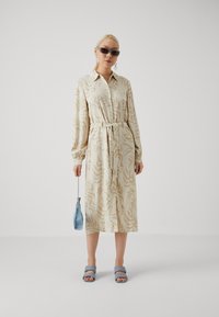 Robe de couleur crème avec des motifs abstraits beiges, manches longues, col boutonné et ceinture à la taille. Portée avec des sandales bleues et un sac à main bleu.
