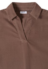 Chemise en velours côtelé marron avec un col en V et une étiquette blanche portant la mention "CECIL" à l'intérieur de l'encolure.