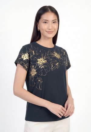 Junge Frau, die ein schwarzes kurzärmeliges Shirt mit goldenen Blumenmustern trägt und mit locker verschränkten Händen vor einem weißen Hintergrund steht.