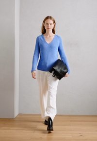 Maglione azzurro lavorato a maglia con scollo a V, pantaloni bianchi a pieghe e borsa in pelle nera. Modella che indossa stivaletti neri alla caviglia. Sfondo grigio uniforme.