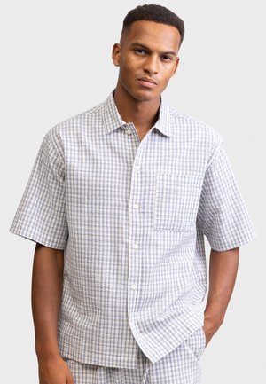 Joven con camisa de manga corta abotonada con patrón de cuadros en azul claro y blanco, posando frente a un fondo gris liso.