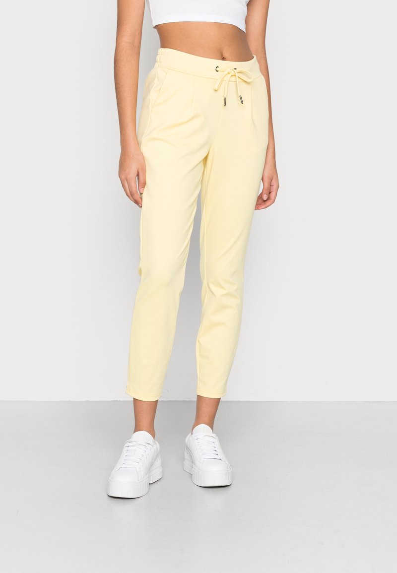 Femme portant un pantalon jaune pâle à cordon de serrage et des baskets blanches à plateforme, debout devant un fond clair uni.