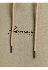 Beige hættetrøje stof med to snore og sort tekst "Premium EST 1990" trykt på brystområdet.