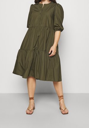Kobieta w oliwkowozielonej sukience midi z warstwami, z bufkami o długości do łokcia oraz brązowych sandałach na słupku z paskami.