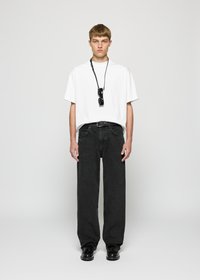 Mann som står mot en enkel bakgrunn iført en hvit oversized t-skjorte, løse sorte jeans, svarte sko og svarte solbriller hengende på en snor rundt halsen.