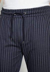 Detailní záběr na tmavě modré kalhoty s tenkými bílými svislými proužky a staženou černou šňůrkou v pase.