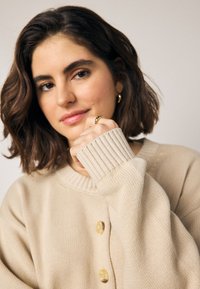 Pull en maille beige avec poignets côtelés et encolure ronde. Comprend trois gros boutons ronds au centre. Texture douce, coupe décontractée.