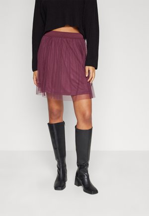 Femme portant une jupe en tulle prune, un pull court noir et des bottes noires à talons montant jusqu'aux genoux, debout sur un fond blanc uni.
