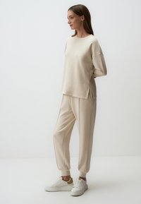 Beige gebreide sweatshirt met verlaagde schouders, zijslits; bijpassende taps toelopende broeken met elastische boorden; gecombineerd met witte sneakers.
