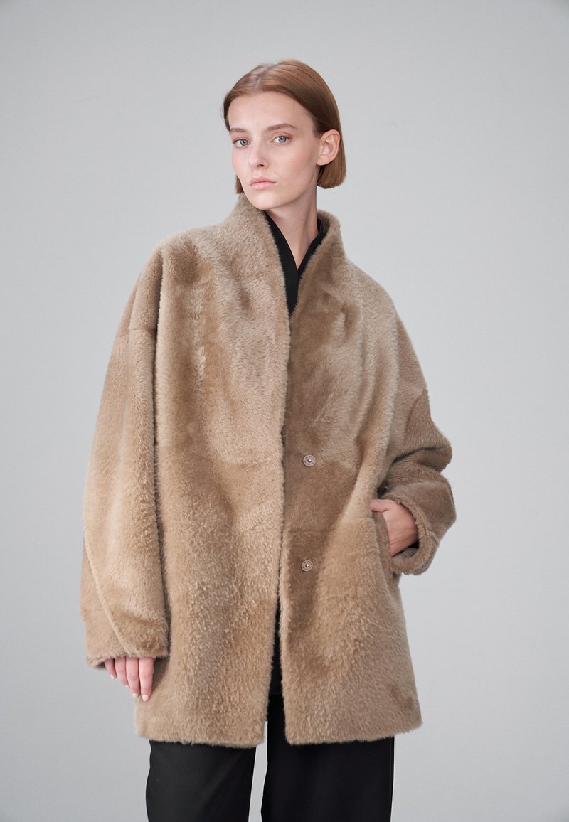 Palton bej oversized din blană artificială, cu o textură moale, guler mare și două butoane frontale, având buzunare laterale și o croială lejeră.