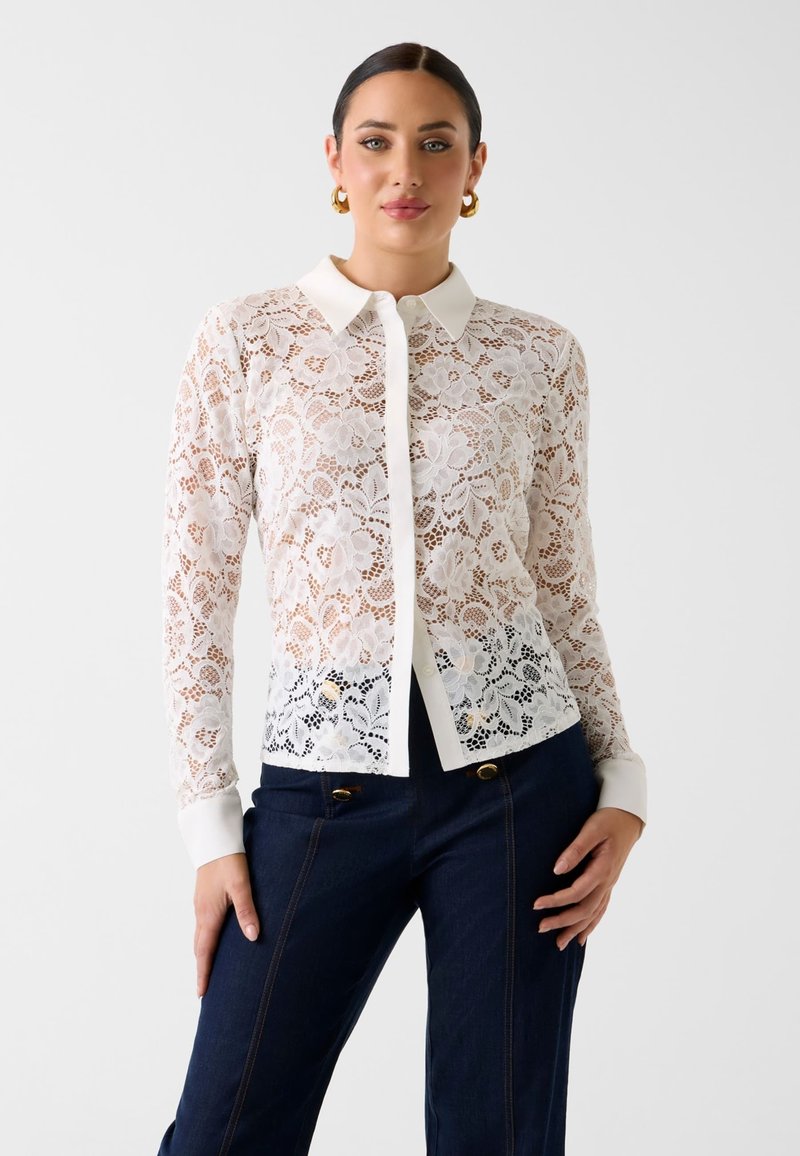 Femme aux cheveux foncés portant une chemise blanche en dentelle fleurie boutonnée et un pantalon bleu foncé, posant avec une main sur la hanche devant un fond uni.