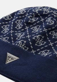 Gorro de punto en azul marino con patrón geométrico blanco. Presenta una etiqueta triangular de logo plateado en la parte frontal. Tejido suave y texturizado.