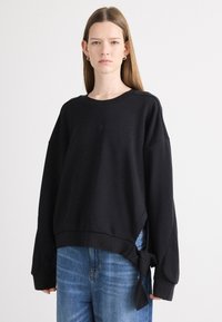 Felpa nera oversize con maniche lunghe, caratterizzata da un dettaglio a nodo sull'orlo. Realizzata in morbido tessuto con scollo a girocollo e logo discreto.