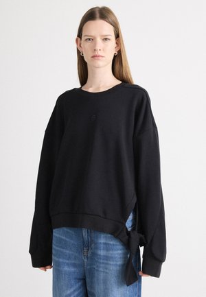 Crni oversized dukserica s dugim rukavima, s detaljem čvora na rubu. Izrađena od mekanog materijala s okruglim izrezom i suptilnim logotipom.