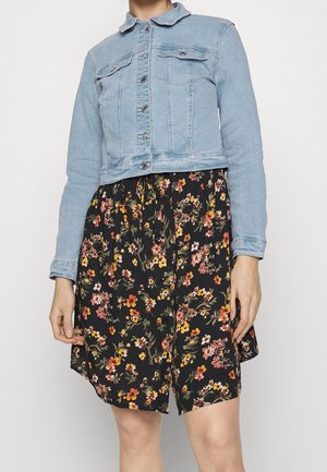 Personne portant une veste en jean bleu clair sur une jupe noire à motifs floraux avec des fleurs orange, jaunes et roses, debout devant un fond uni.