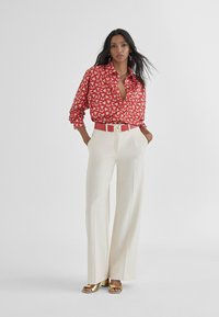 Blusa rossa a fantasia con maniche lunghe abbinata a pantaloni a gamba larga color crema e una cintura coordinata con un logo prominente. Sandali con tacco dorati.