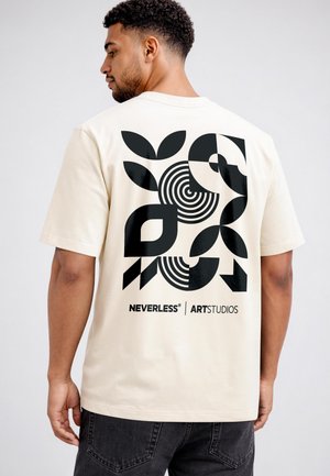Man in light beige t-shirt with bold black geometric shapes and NEVERLESS ART STUDIOS text on the back, wearing dark jeans.