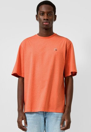 Młody mężczyzna stoi, ubrany w pomarańczową koszulkę z napisem "THIS IS QS" na piersi i jasnoniebieskie dżinsy, na jednolitym jasnym tle.