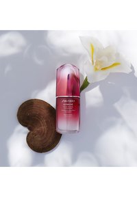 Shiseido Ultimune-serum i en gradient pink glasflaske, med en afrundet top og slankt design, placeret på en hjerteformet træskive ved siden af en hvid blomst.