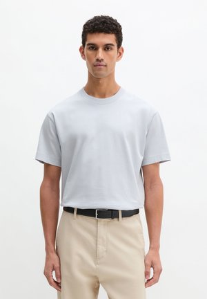 Mężczyzna z krótkimi kręconymi włosami, ubrany w jasnoszarą koszulkę z okrągłym dekoltem, włożoną w beżowe spodnie z czarnym paskiem, stojący na białym tle.