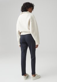 Maglione bianco e soffice con maniche ampie, abbinato a pantaloni navy su misura e scarpe da ginnastica di colore chiaro. Sfondo neutro.
