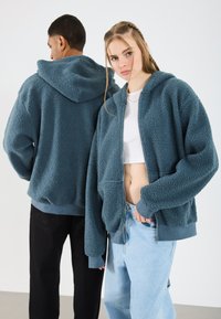 Fleece zip-up hoodie in teal, met een gestructureerde buitenkant, voorzakken en een trekkoord capuchon. Gecombineerd met losse lichtblauwe jeans.