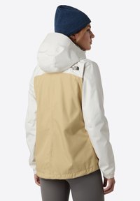 Damjacka med vit överdel och beige underdel, tillverkad av ett lätt material, med huva och elastiska ärmslut.