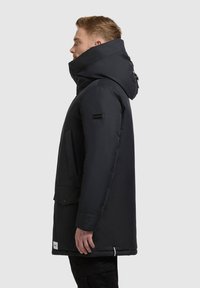 Melnā kapucēta parka ar spilvenu dizainu, spiedpogu aizdari un logotipa uzlīmi uz piedurknes. Iespējas ietver divas lielas priekšējās kabatas.