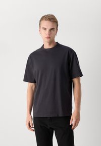 Camiseta negra de manga corta con cuello redondo y acabado texturizado, que presenta un logo sutil en el escote y un corte relajado.