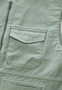 Mintfärgad väst i textil med struktur, med dragkedjestängning och en bröstficka med klaff. Sömnadsdetaljer synliga.