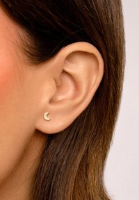 Goldene Halbmondohrringe mit kleinen klaren Edelsteinen, die am Ohrläppchen getragen werden. Haare verdecken teilweise das Ohr. Geschmeidige Hauttextur sichtbar.