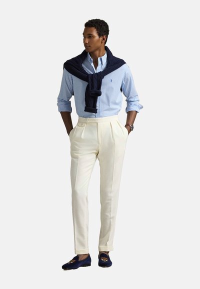 Chemise à boutons bleu rayée, pantalon plissé blanc, et pull marine drapé sur les épaules. Chaussures navy avec des détails brodés.