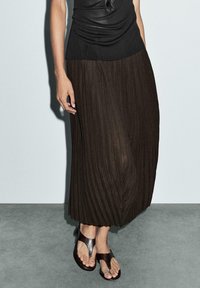 Donna che indossa un top senza maniche nero, una gonna midi plissettata marrone e sandali in pelle nera con fascia tra le dita, in piedi su un pavimento grigio contro un muro bianco.